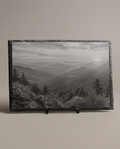 A rectangular black slate plaque featuring a black and white landscape photograph of misty mountain ranges and pine trees, displayed on a small black stand against a neutral beige background.